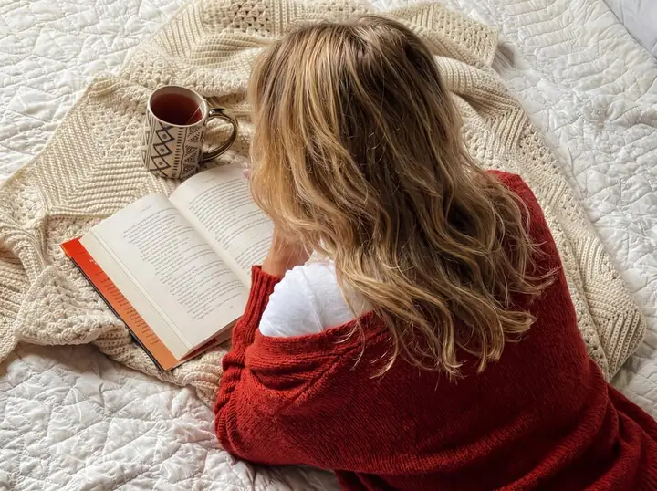 Woman in a red sweater lying on her bed with a book and a cup of tea